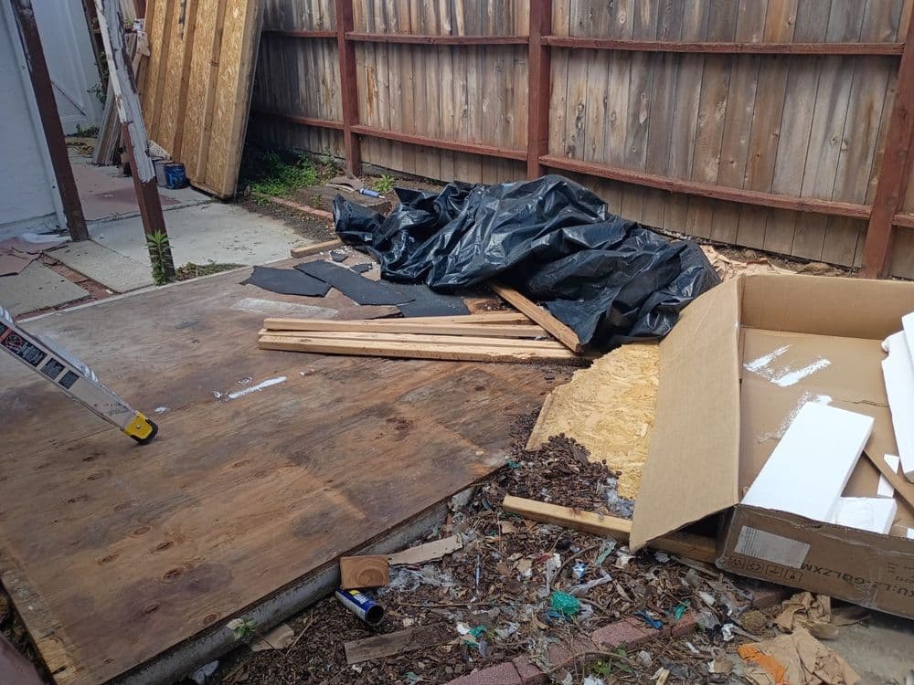 Construction debris in messy backyard with wooden planks, black tarp, and cardboard box.