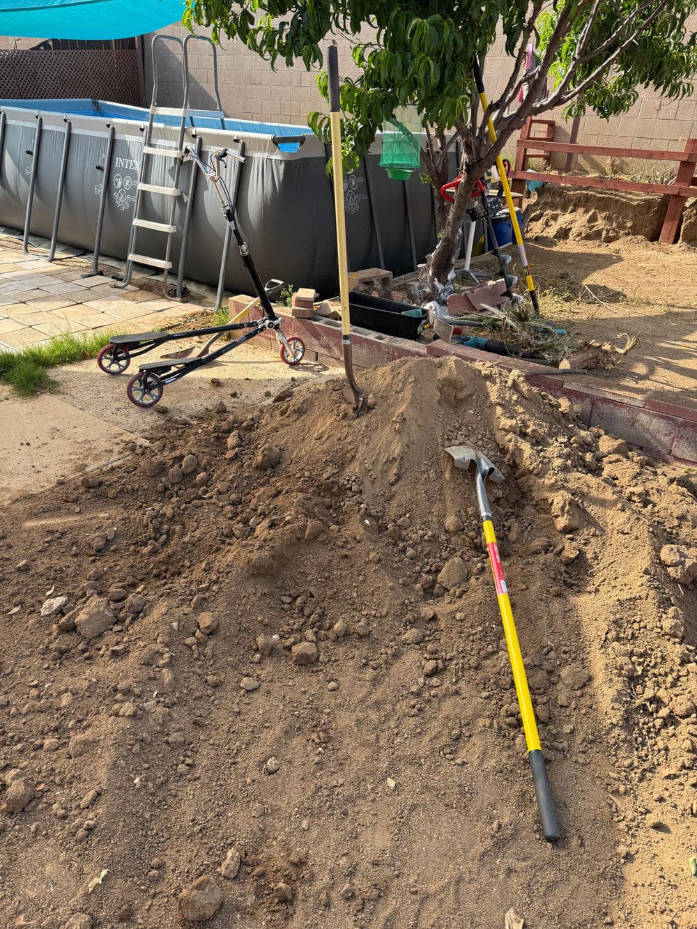 Dirt pile with gardening tools, scooters, and an above-ground pool in a backyard setting.
